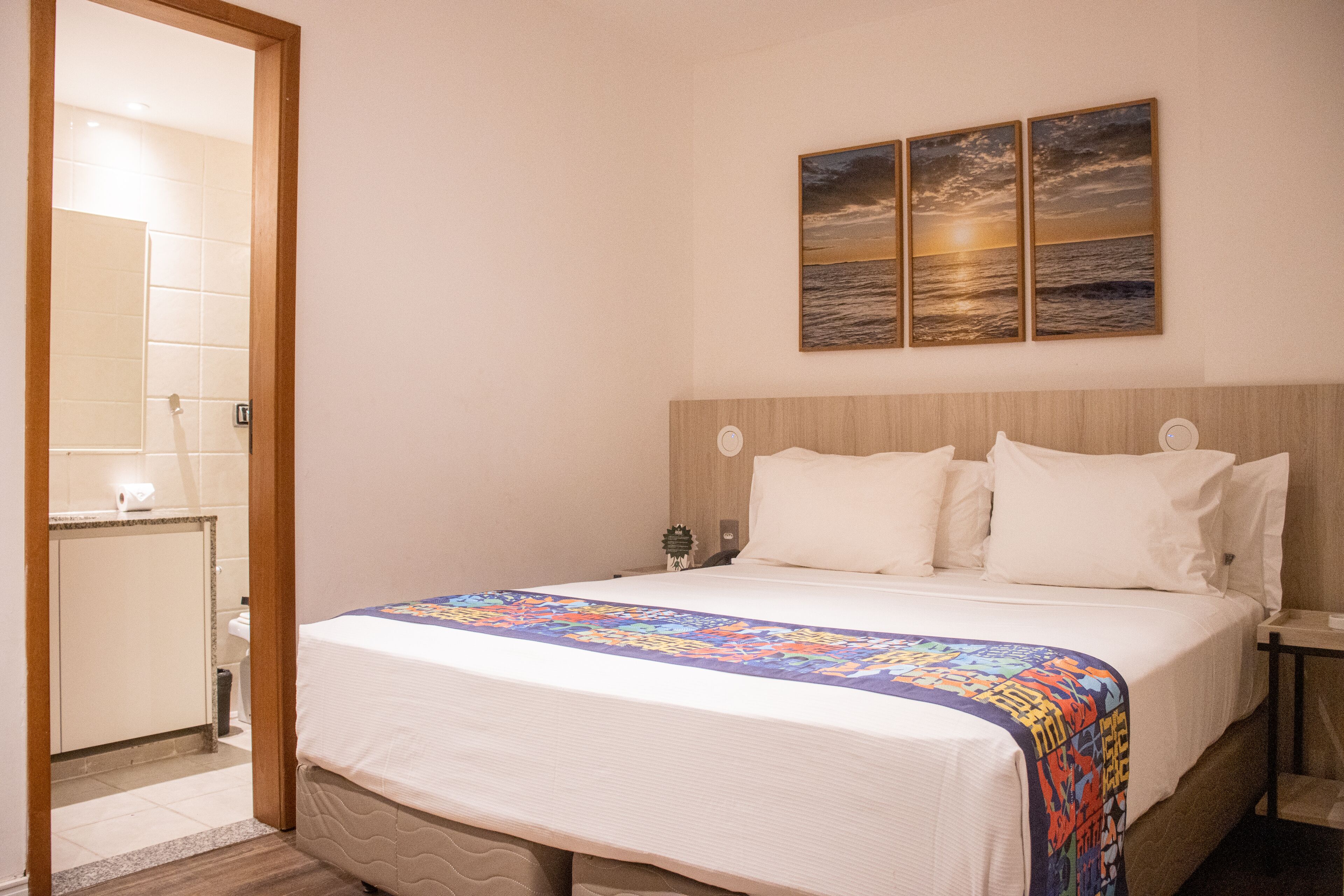 Een hotelkamer met een groot bed, een houten hoofdeinde, een nachtkastje en een schilderij van een zonsondergang boven de oceaan.
