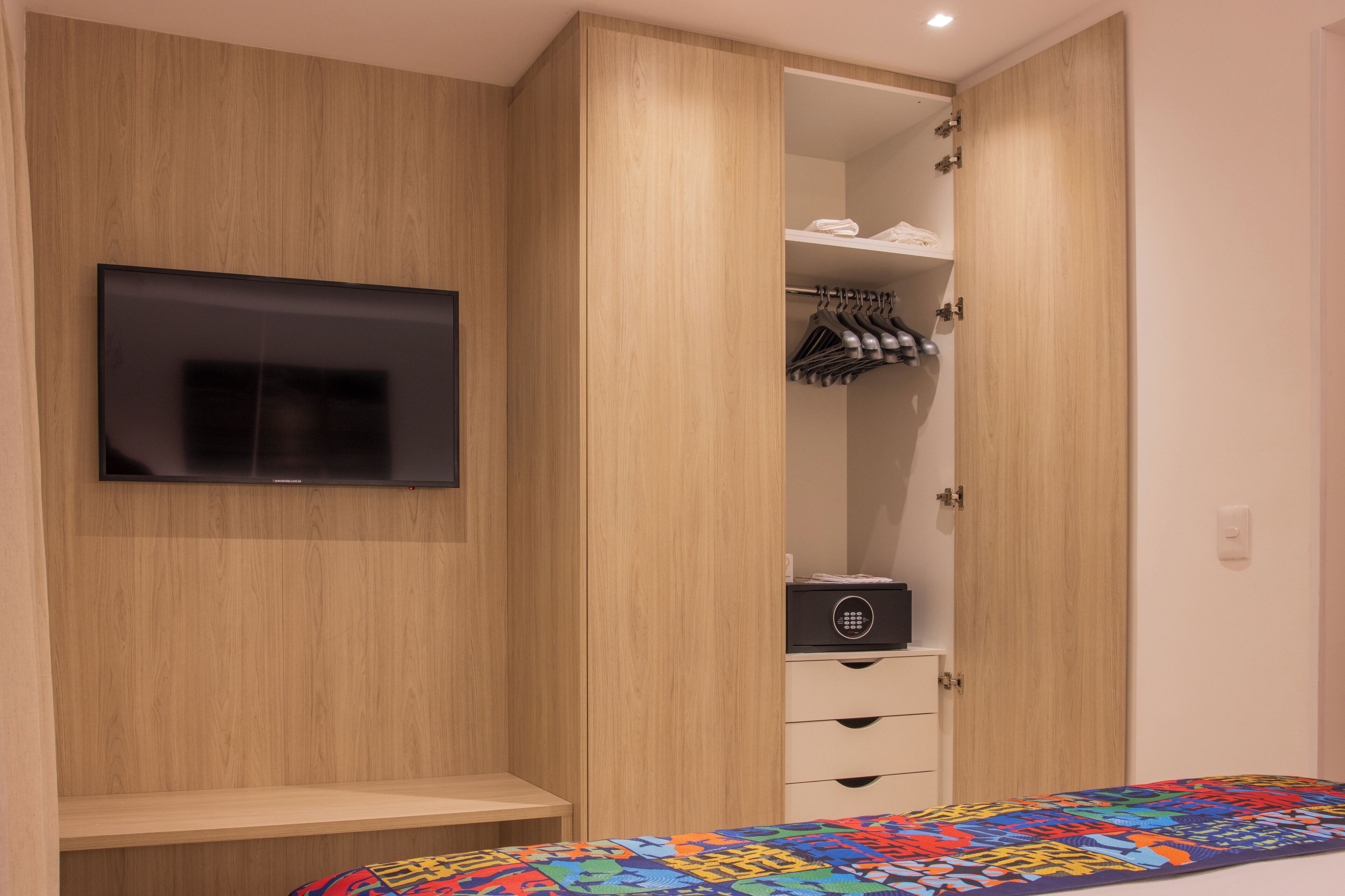 A bedroom with a bed, a TV mounted on the wall, a wooden wardrobe, and a bedside table.