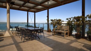Un espacio al aire libre techado con una mesa y sillas de madera, plantas en macetas y vista al mar.