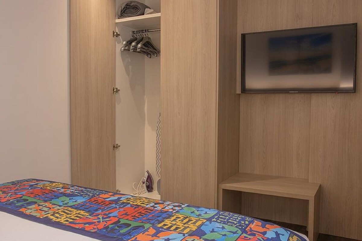 A bedroom with a bed, a TV mounted on the wall, and a wardrobe with hanging clothes.