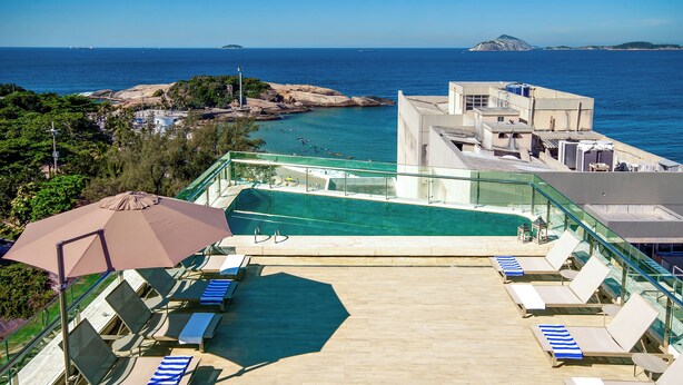 Zona de piscina en la azotea con sillones y sombrillas, con vistas a un paisaje costero de aguas azul turquesa y afloramientos rocosos.