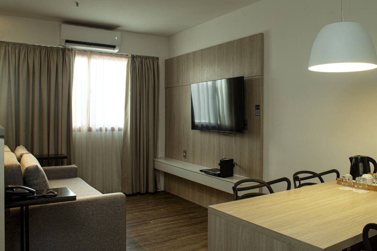 A modern hotel room with a sofa, a dining table, chairs, a TV, and a coffee maker.