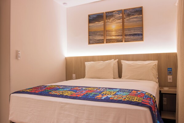 A hotel room with a bed, a colorful quilt, a wooden headboard, and a sunset painting on the wall.