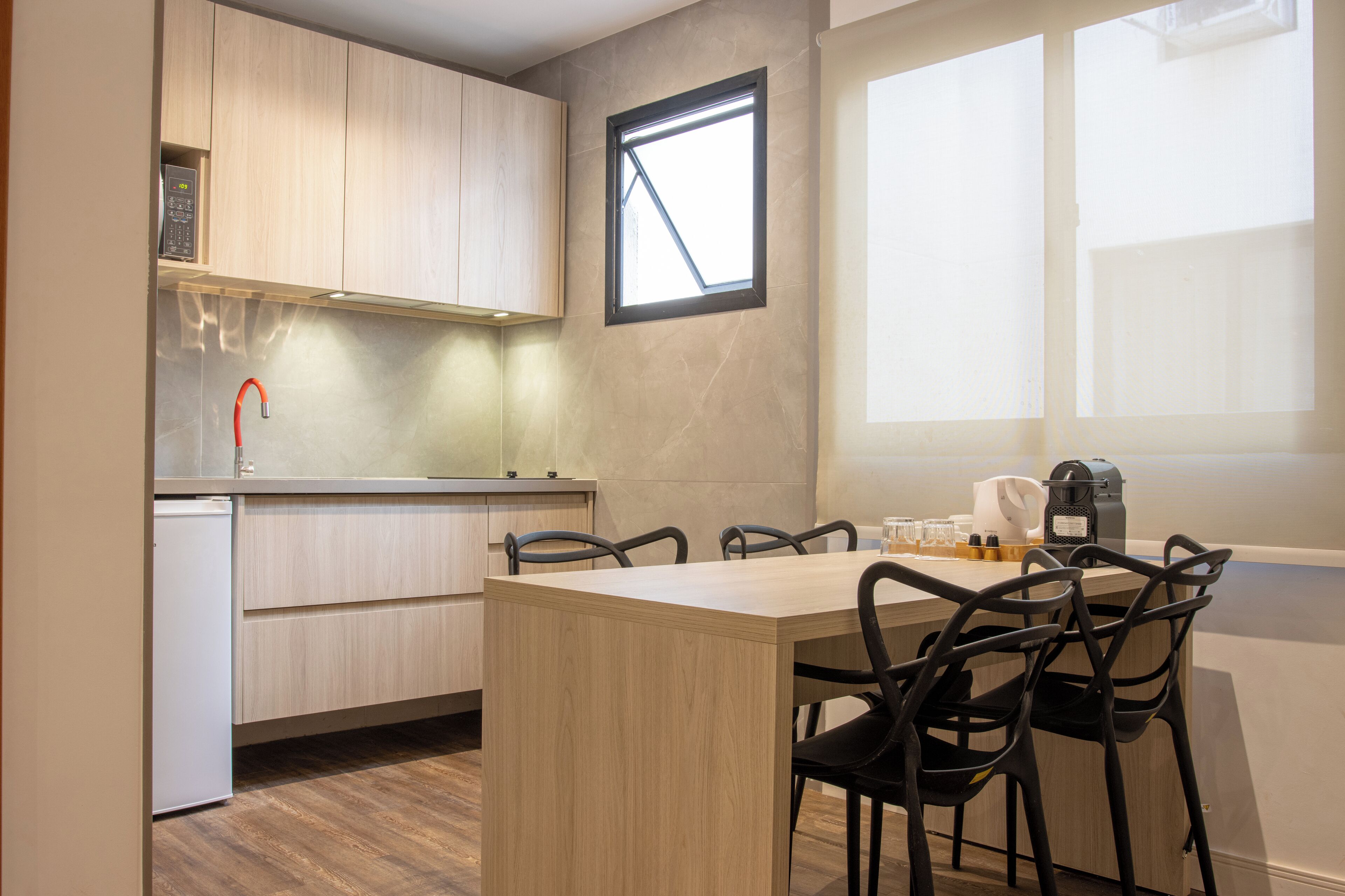 A modern kitchen with wooden cabinets, a built-in oven, a coffee machine, and a dining area with a table and chairs.