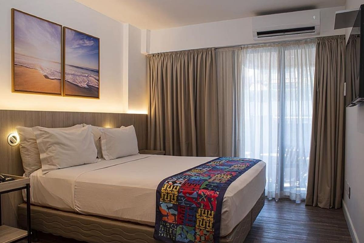 A hotel room with a bed, a nightstand, a wall-mounted TV, and a window with curtains.