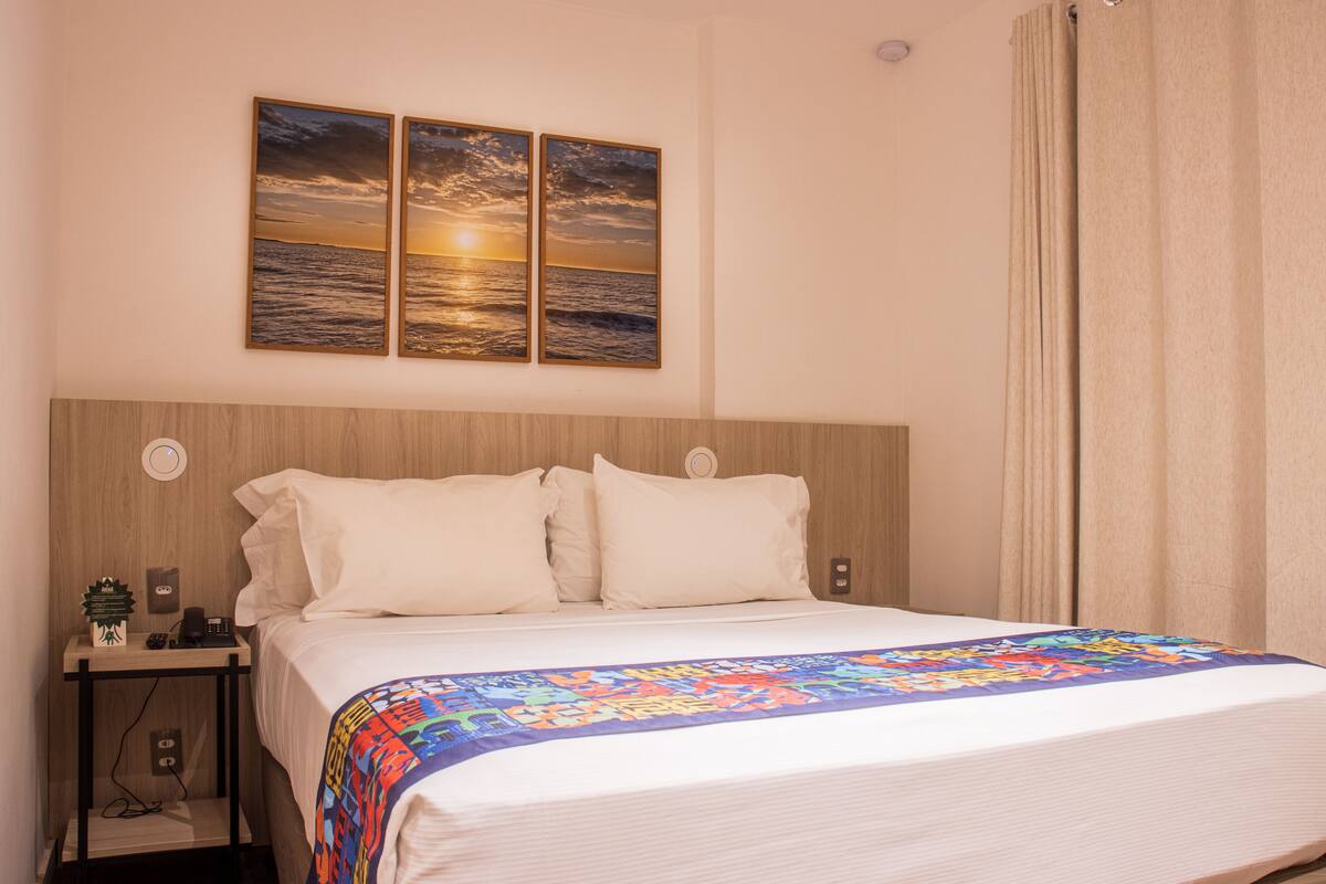 A hotel room with a bed, a nightstand, a phone, and a wall-mounted artwork depicting a sunset over the ocean.