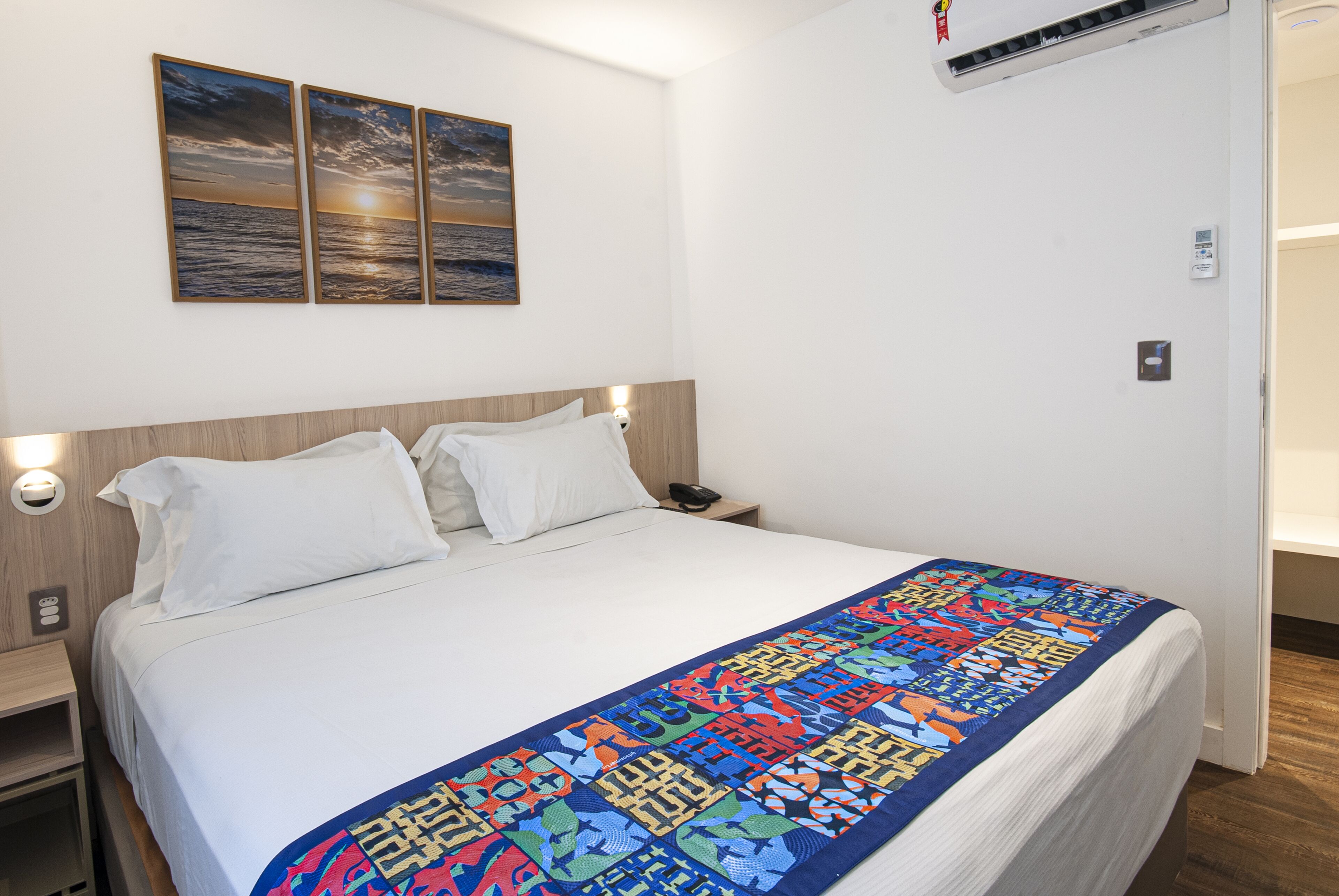 A neatly made bed with white linens and a colorful blanket, a wooden headboard, and a wall-mounted air conditioner.
