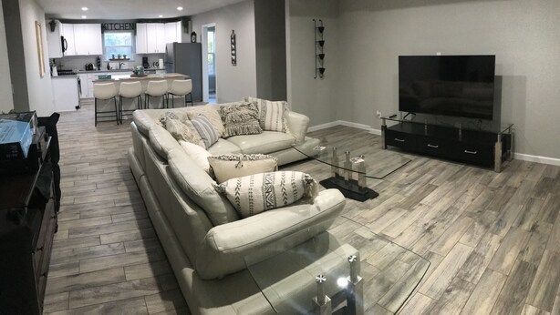 Een moderne woonkamer met een bank, televisie en een glazen salontafel. Achtergrond toont een keuken met witte kasten en een eethoek met een tafel en stoelen.