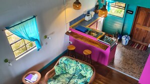 A cozy room with wicker furniture, a kitchen area, and a window with blue curtains. - BLUE COTTAGE Almost Paradise (Sauteurs)