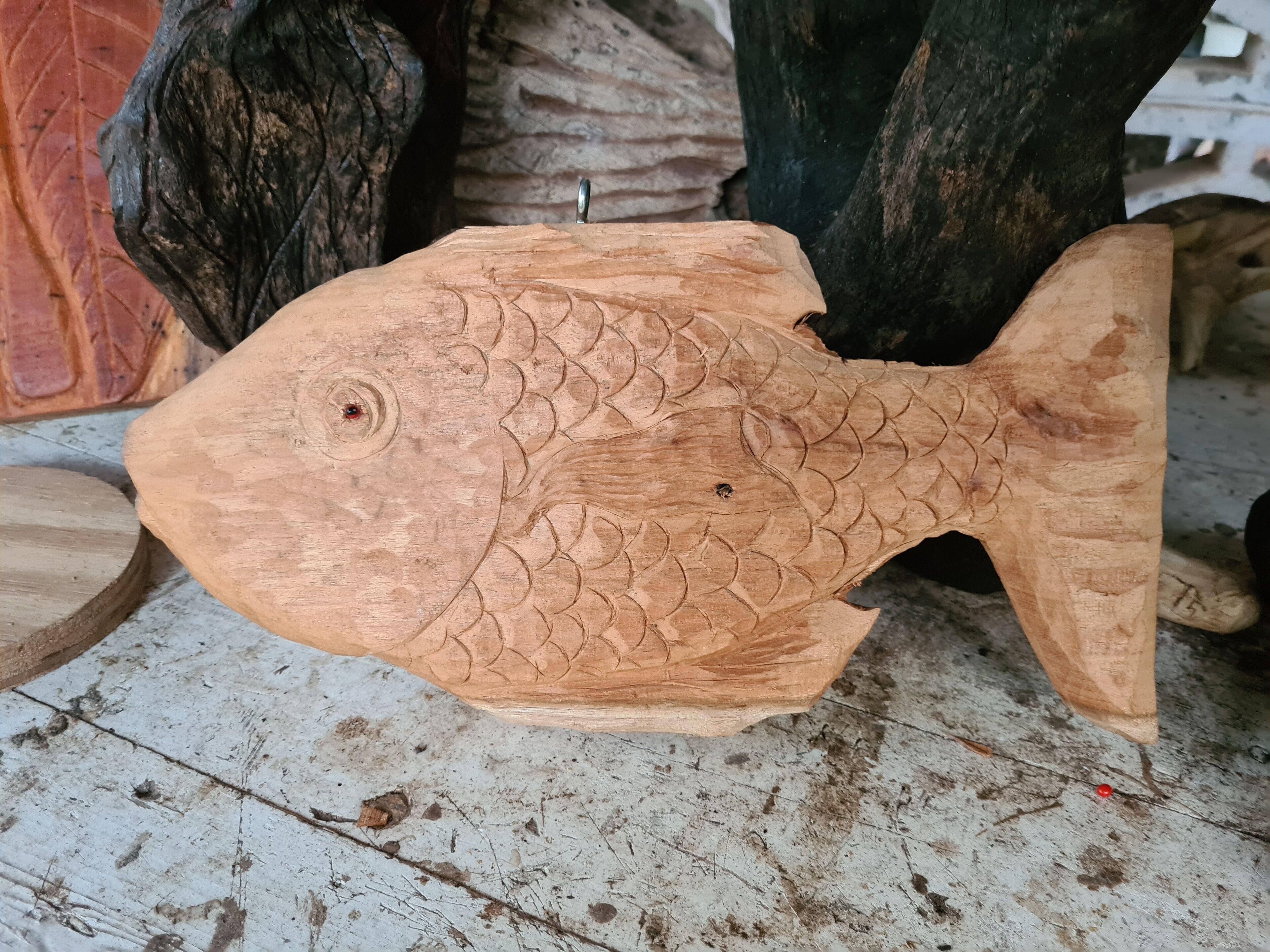 A wooden fish sculpture with detailed scales and fins, placed on a concrete floor with other wooden objects nearby.