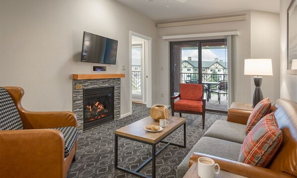 A living room with a fireplace, a television mounted above it, and a balcony with outdoor seating.