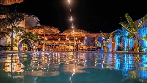 A nighttime pool area with clear water reflecting lights, surrounded by palm trees and umbrellas.