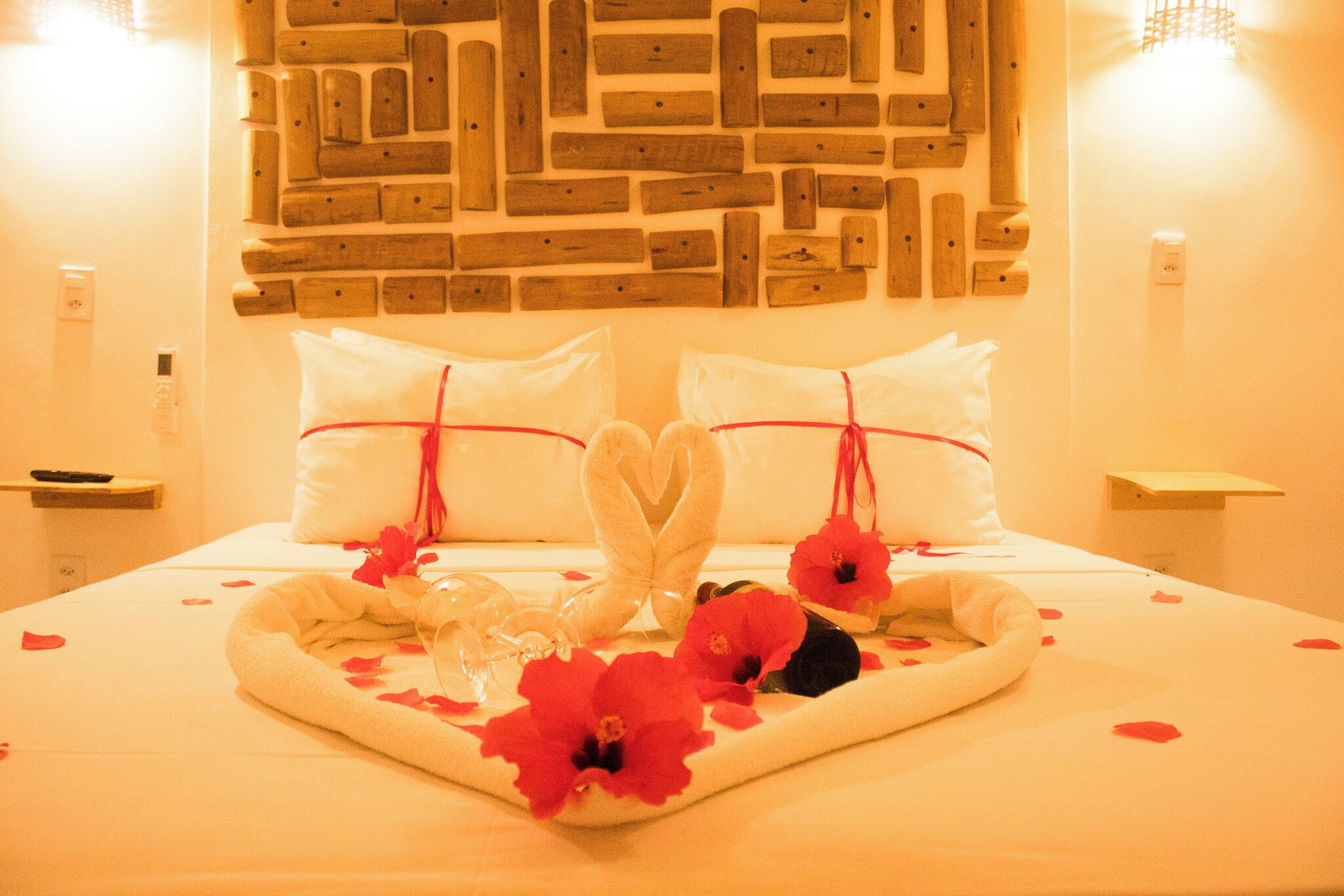 A neatly made bed with heart-shaped pillows, a tray with rose petals and a glass, and a wooden wall decor.