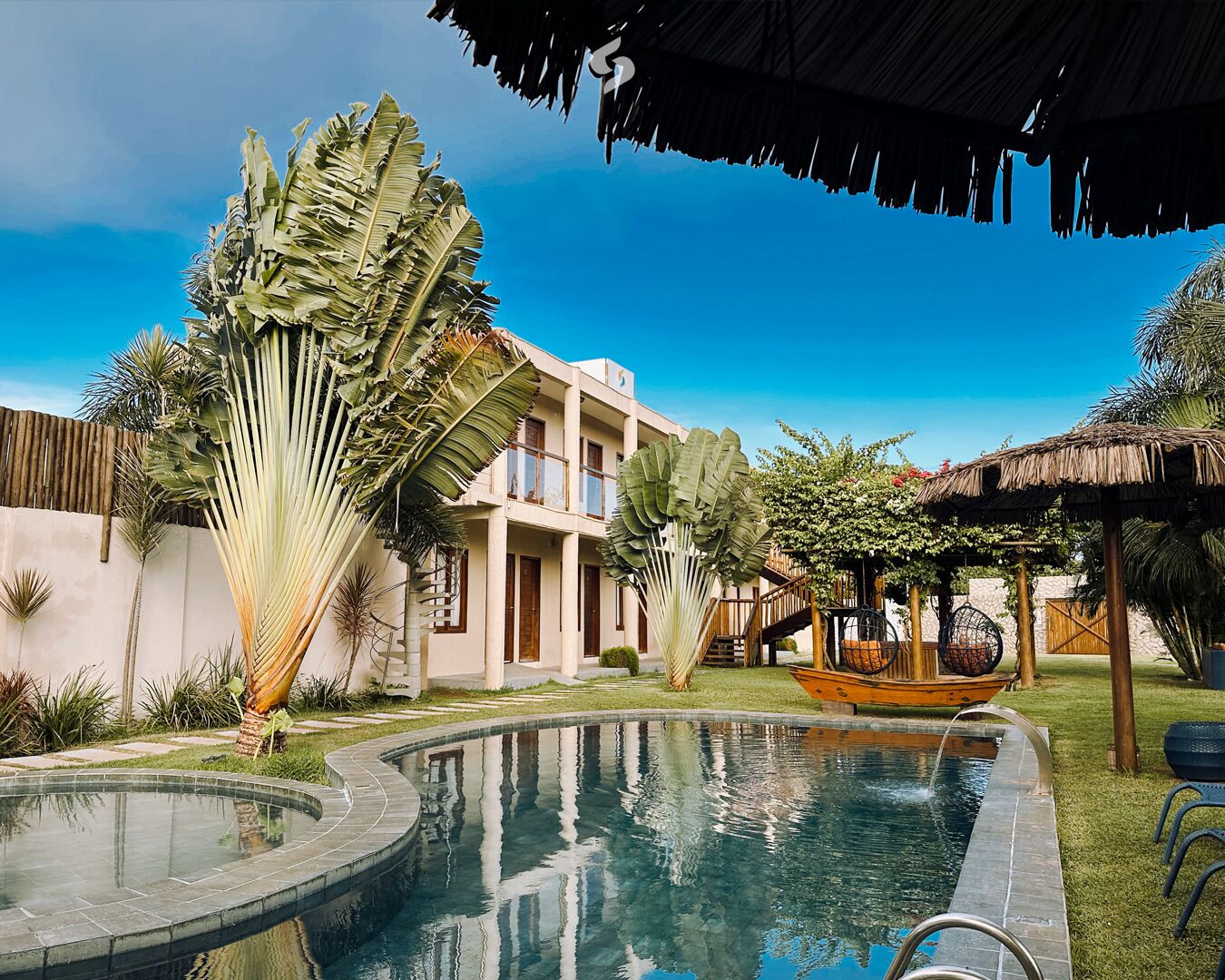 A pool with a wooden deck, a thatched-roof structure, and a multi-story building with balconies.