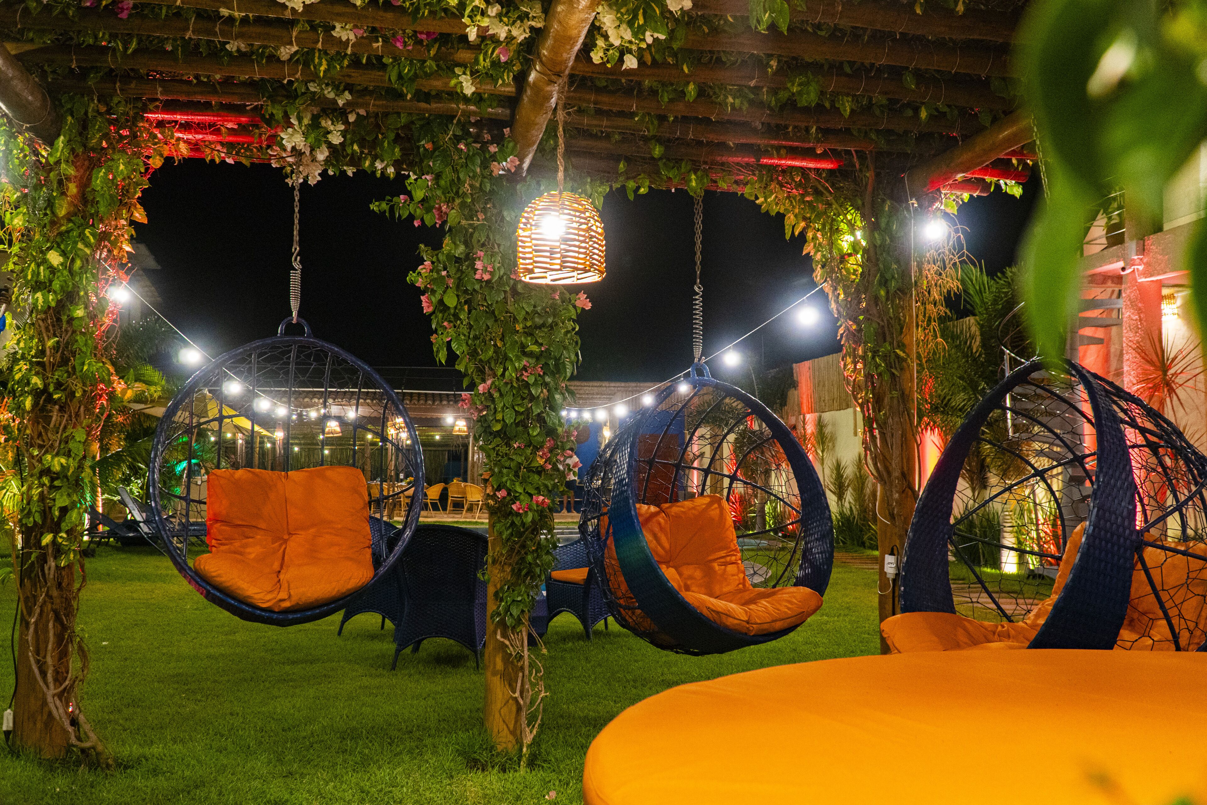Un espacio de estar al aire libre con sillones colgantes y una mesa, rodeado de follaje y luces colgantes.