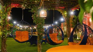 Un espacio de estar al aire libre con sillones colgantes y una mesa, rodeado de follaje y luces colgantes.