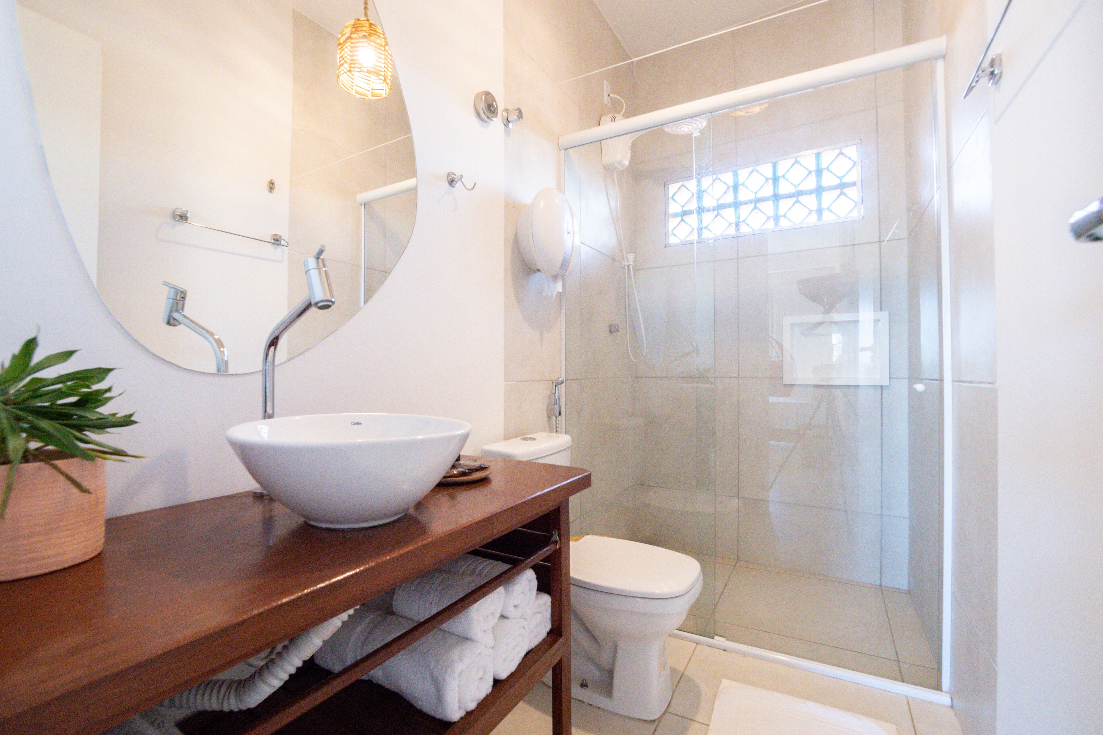 Un baño moderno con un espejo grande, un lavamanos blanco, ducha con mampara de vidrio y una planta en maceta.