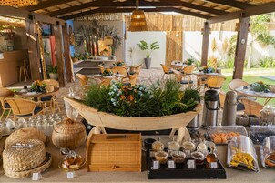 Uma área de refeições ao ar livre com mobiliário de vime, um arranjo de plantas no centro e diversos recipientes com comida e bebida.
