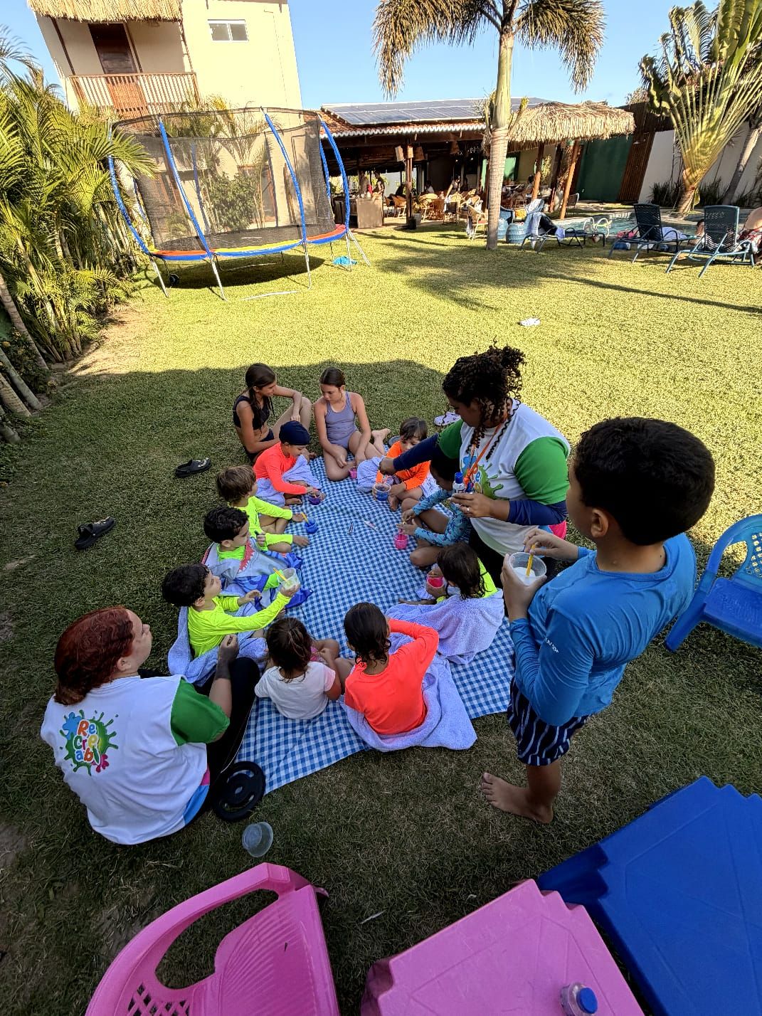 A group of children and adults sitting on a checkered mat outdoors, engaged in an activity.