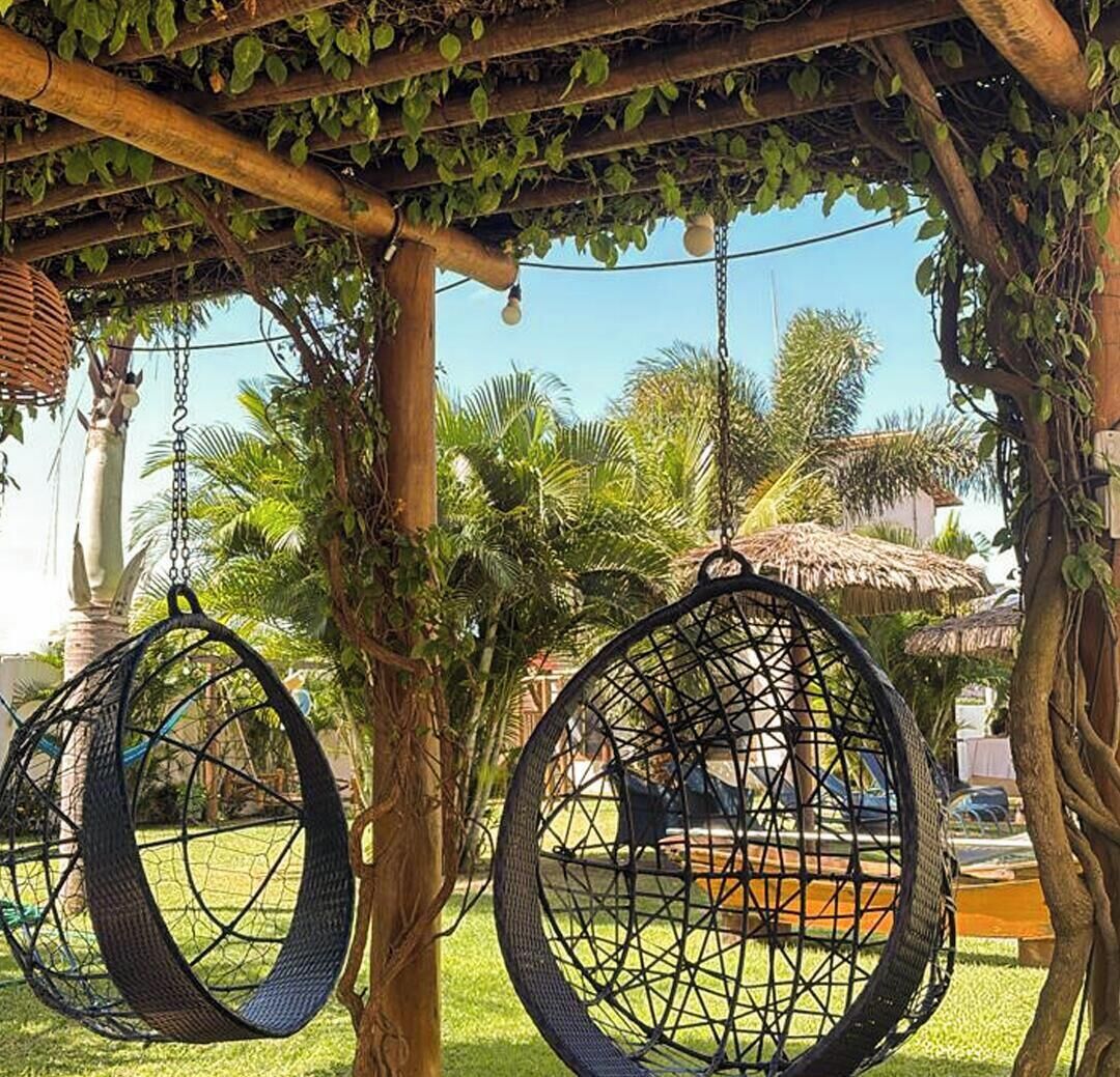 A hammock swing under a wooden pergola with greenery, a thatched umbrella, and a palm tree in the background.