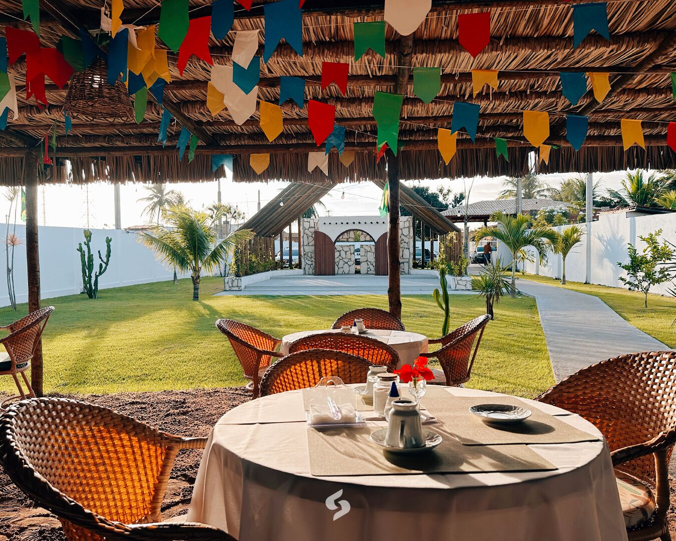 An outdoor dining area with wicker chairs and a table set for two, under a thatched roof with colorful flags.