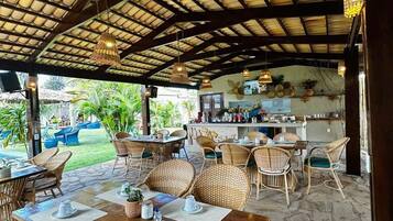 Un área de comedor al aire libre con mobiliario de mimbre, techo cubierto y una cocina al fondo.