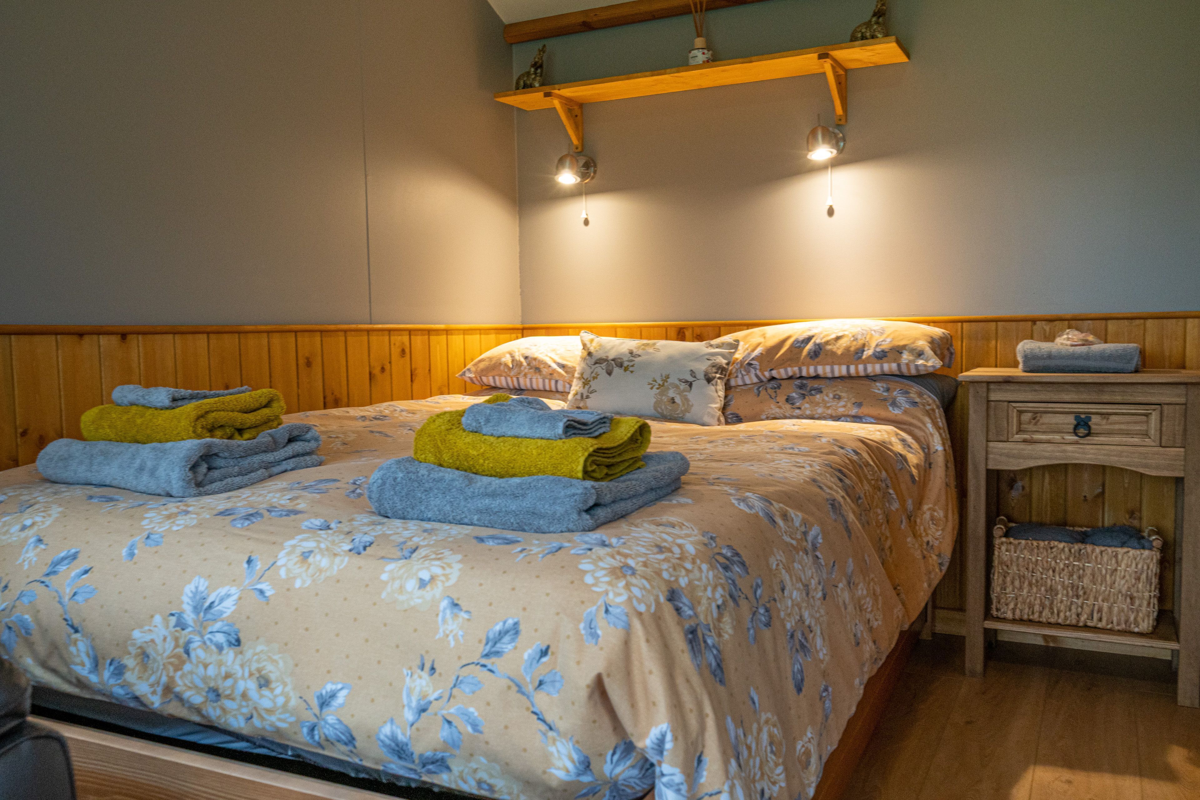 Une chambre avec une tête de lit en bois, un lit recouvert d’une literie à motifs floraux, une table de chevet avec une lampe et une petite table de nuit en bois avec un panier.