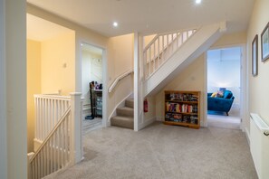 Un couloir recouvert de moquette, avec un escalier blanc, une bibliothèque et un canapé bleu en arrière-plan.