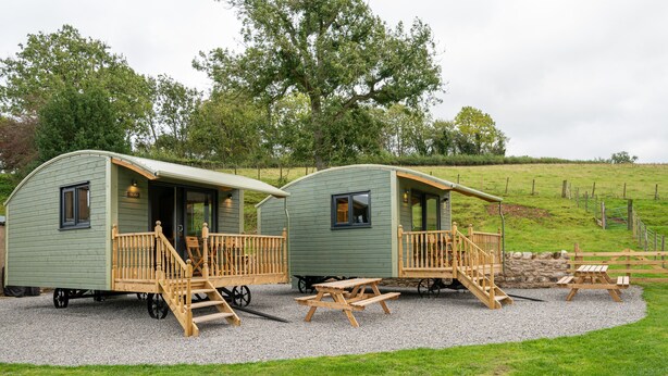 Two green mobile homes with wooden porches and picnic tables, set on a gravel area with a grassy field in the background.