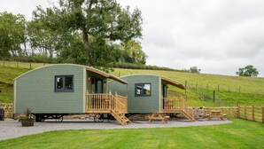 Dos casas móviles verdes con balcones de madera y una mesa de picnic, ubicadas en una colina cubierta de césped con árboles al fondo.