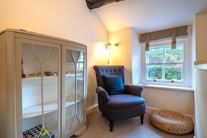 A room with a large glass cabinet, a tufted armchair with a blue pillow, a woven basket, and a window with a view of greenery.