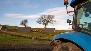A blue tractor with a cabin and a mounted orange light, set against a rural landscape with green fields and a clear sky.