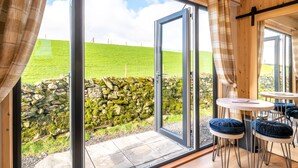 Ein Zimmer mit einer großen Glasdoor, die zu einer Steinmauer und einem grasbewachsenen Hügel führt. Es gibt zwei runde Tische mit Stühlen.