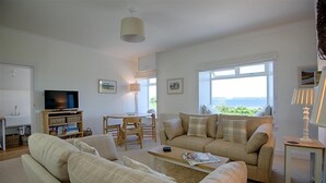 A spacious living room with a sofa set, a television, a dining table with chairs, and large windows offering a view of the outdoors.