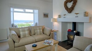 A living room with a beige sofa, a wooden coffee table, a fireplace with a heart-shaped decoration, and a view of the sea through a large window.