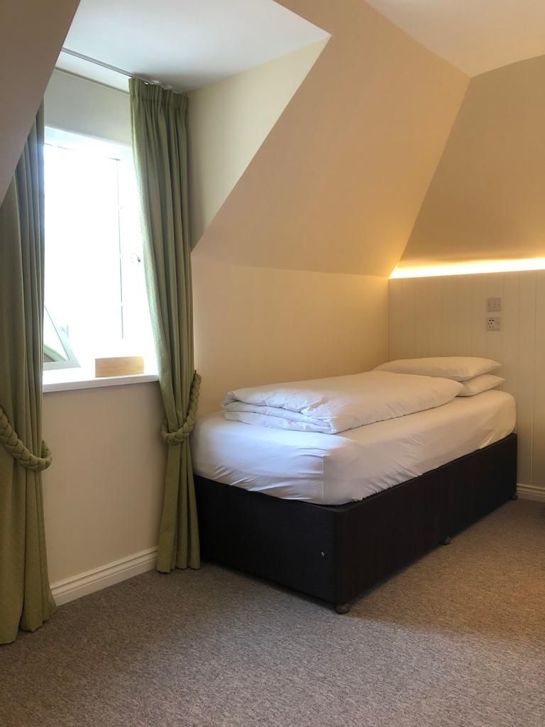 Une petite chambre avec un lit simple, un plafond en pente, une fenêtre avec des rideaux et un luminaire fixé au mur.