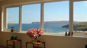 A room with a large window overlooking a coastal landscape, a vase with flowers, and a small table with a green object. - Sangobeag -  a cottage that sleeps 6 guests  in 3 bedrooms (Durness)