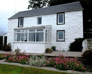 A two-story white house with a large bay window, surrounded by a garden with pink flowers. - Swallow Cottage - a cottage that sleeps 6 guests in 3 bedrooms (Dalbeattie)