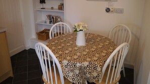 Ein Essbereich mit einem runden Tisch, bedeckt mit einer gemusterten Tischdecke, umgeben von weißen Stühlen. Auf dem Tisch steht eine Vase mit Blumen.