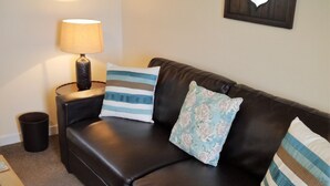 Ein dunkelbraunes Ledersofa mit drei Kissen, ein kleiner Tisch mit einer Lampe, ein gerahmtes Bild an der Wand und ein Mülleimer.