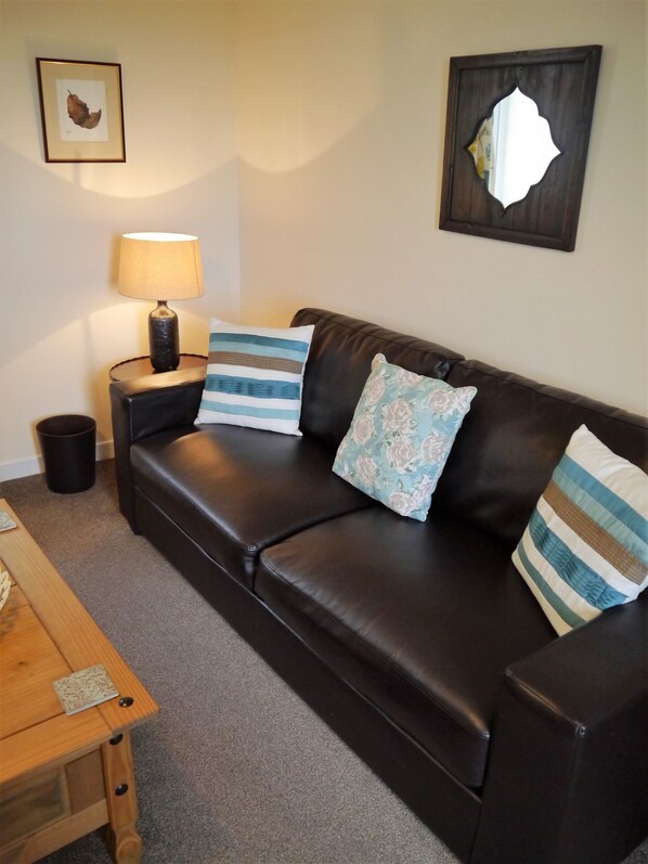A dark brown leather sofa with three pillows, a small table with a lamp, a framed picture on the wall, and a trash can.