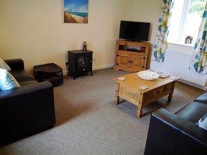 A living room with a sofa, a wooden coffee table, a television on a wooden stand, a small black stove, and a window with curtains.