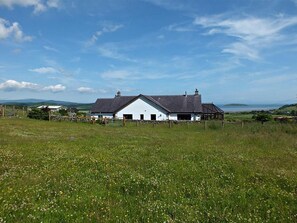 A white building with a dark roof, surrounded by a grassy field with wildflowers, under a clear sky.