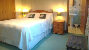 Une chambre à coucher bien rangée, avec un lit, des lampes de chevet, une table de nuit en bois, une petite table et une salle de bain visible à travers une porte ouverte.