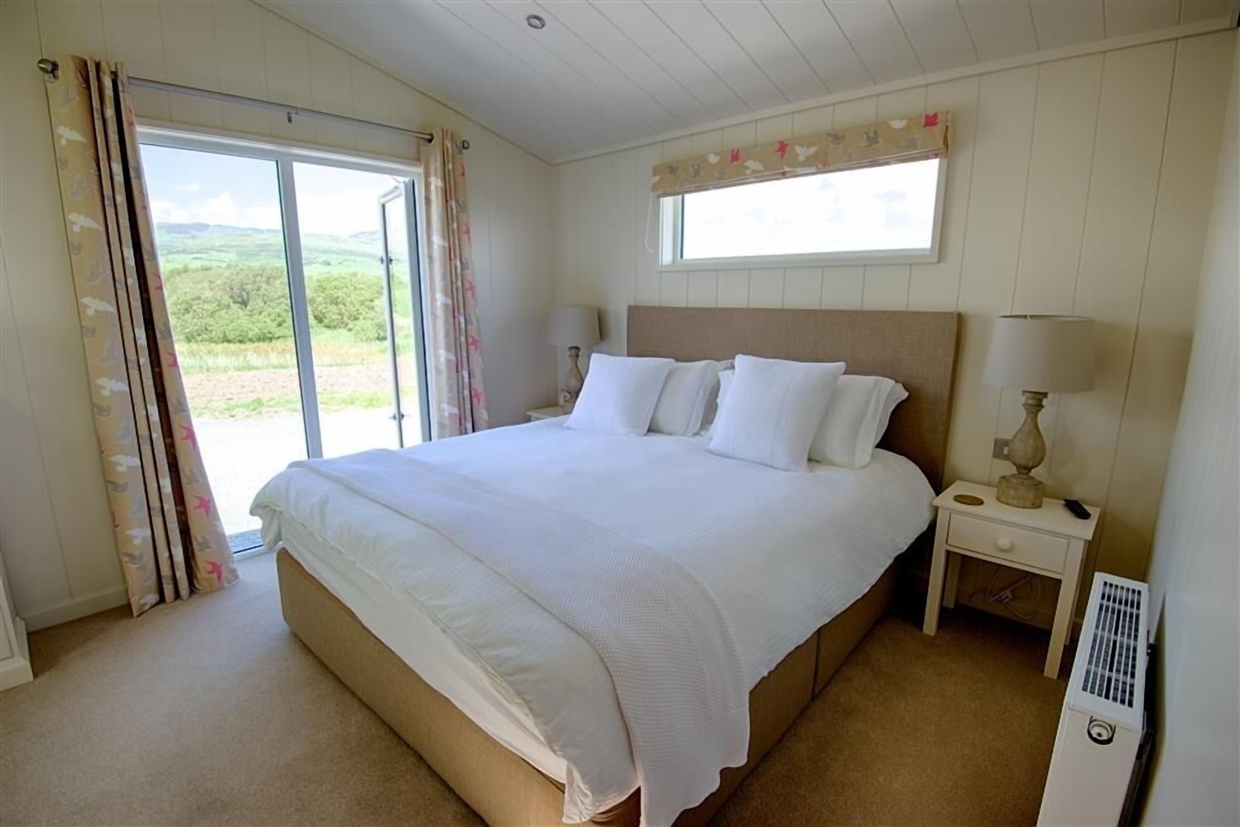 A bedroom with a large bed, a nightstand with a lamp, a window with a view of greenery, and a radiator.