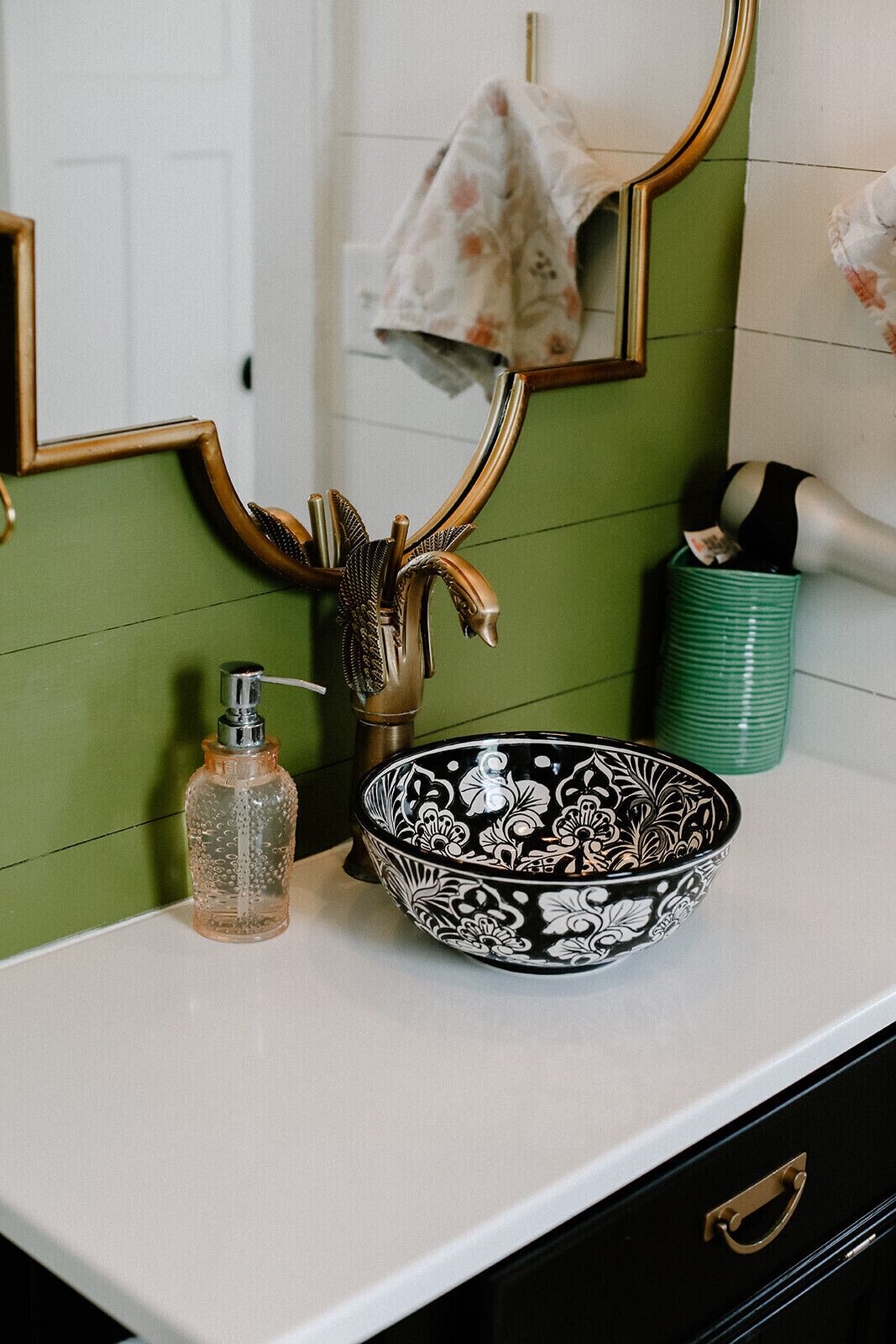 Un tocador de baño con pared verde, un espejo con marco dorado, un cuenco blanco y negro estampado, un dispensador de jabón transparente y un cubo de basura verde.