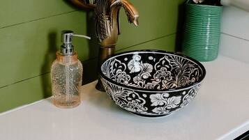 Un tocador de baño con pared verde, un espejo con marco dorado, un cuenco blanco y negro estampado, un dispensador de jabón transparente y un cubo de basura verde.