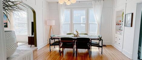 Una habitación amplia con piso de madera, una mesa de comedor con sillas, una araña y ventanas grandes con cortinas traslúcidas.