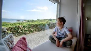 A person sitting on a cushioned bench by a large window overlooking a coastal landscape with greenery and a stone wall. - Threaby Cottage -  a cottage that sleeps 4 guests  in 2 bedrooms (Gatehouse of Fleet)