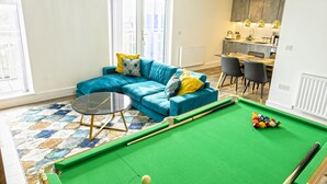 A living room with a pool table, a blue sofa, and a dining area with chairs and a kitchen in the background.